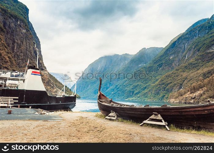Old wooden viking boat and ferry boat on seashore in norwegian nature, foggy misty day. Mountains and fjord Sognefjord. Tourism and traveling concept. Old wooden viking boat in norwegian nature