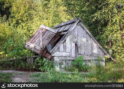 Old wooden well with a roof and open door where stands the metal bucket with a chain on a background of foliage in the sunlight.. Old Wooden Well With A Roof