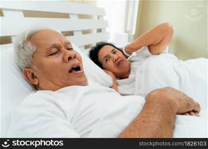 Older Asian woman laying open her eyes and Cover ears with her hands in bed beside her husband who snores and Makes noise, Marital problems.