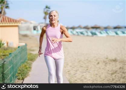 Older female doing sport to keep fit. Mature woman running along the shore of the beach. Concept of healthy living in the elderly. Senior woman in fitness clothing running along beach. Older female doing sport to keep fit. Mature woman running along the shore of the beach.