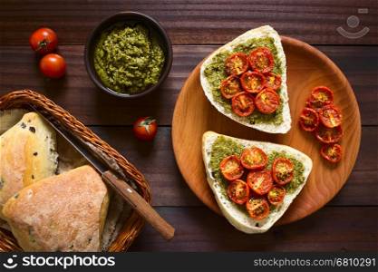 Olive bread roll halves spread with basil pesto and topped with roasted cherry tomato halves, served on wooden plate, photographed overhead on dark wood with natural light