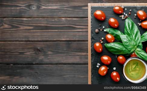 Olive oil and cherry tomatoes with basil leaves, salt and pepper, layout on a black stone board. Ingredients for making Caprese salad. Copy space on a dark wooden background, top view
