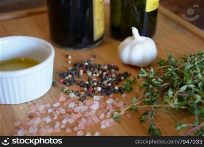 olive oil in a ramekin dish, bottle of olive oil, bottle of balsamic vinegar, garlic, thyme, pink salt, and peppercorns on a wood cutting board