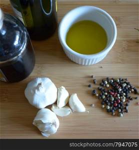 olive oil in a ramekin dish, bottle of olive oil, bottle of balsamic vinegar, garlic, and peppercorns on a wood cutting board