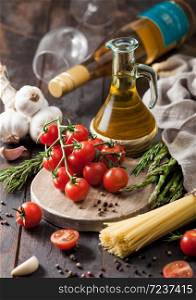 Olive oil with cherry tomatoes on round chopping board and with spaghetti and rosemary with garlic on wooden table background with bottle of white wine and crystal glass.