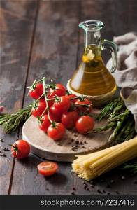 Olive oil with cherry tomatoes on round chopping board and with spaghetti pasta and rosemary and asparagus on wooden table background.