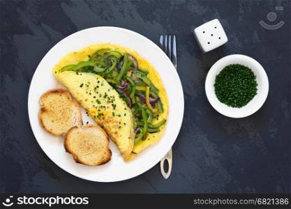 Omelette with green bell pepper and red onion sprinkled with chives, served on plate with toasted and buttered bread slices, photographed overhead on slate with natural light