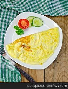 Omelette with slices of tomato, cucumber, parsley on a plate, napkin, fork on the background of wooden boards