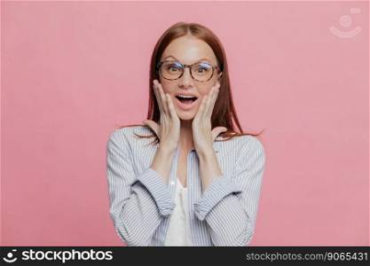 Omg, how nice  Beautiful elegant female employee touches cheeks with both palms, wears spectacles, elegant shirt, cant believe in her promotion, recieves reward, isolated over pink background
