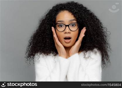 Omg, it cannot be so! Surprised emotional dark skinned young female with curly hair, touches both cheeks, has shocked faciall expression, dressed in white jumper, isolated over grey background