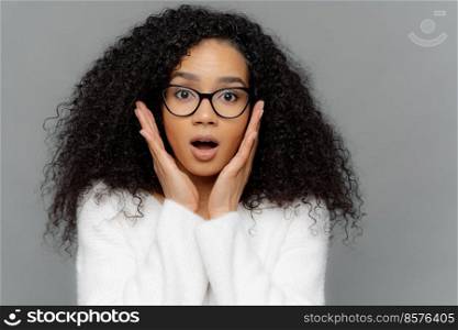Omg, it cannot be so  Surprised emotional dark skinned young female with curly hair, touches both cheeks, has shocked faciall expression, dressed in white jumper, isolated over grey background