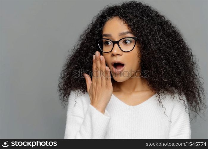 Omg, no way. Scared emotional young dark skinned woman covers opened mouth, feels impressed, has curly hair, wears transparent glasses and white jumper, focused away, isolated on grey background.