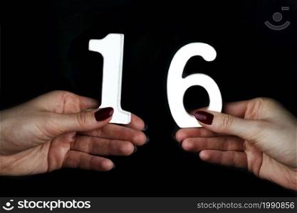On a black background, female hand with number sixteen.. On the female palms figure sixteen.