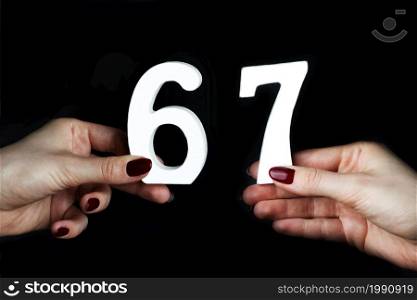 On a black background, female hand with number sixty-seven.. To female hands the number sixty-seven.