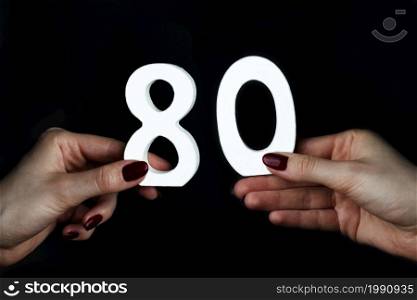 On a black background, female hand with numbers eighty.. To female hands the figure of eighty.