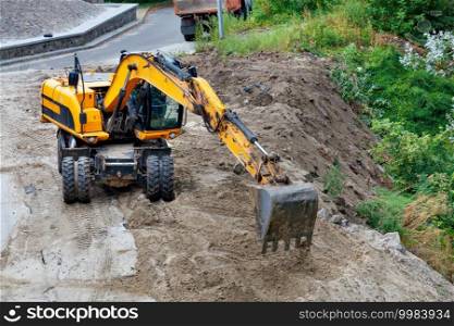 On a cloudy summer day, a construction excavator clears a construction site for the foundation of a future home.. Heavy construction excavator works at a construction site among the near asphalt road.