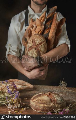On a dark background a baker in a black apron holds a freshly baked bread and wooden table with bread and dried flowers. Baker keeps a variety of bread