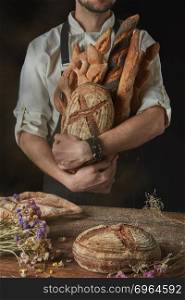 On a dark background a baker in a black apron holds a freshly baked bread and wooden table with bread and dried flowers. Baker keeps a variety of bread