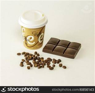 On a gray background is a paper cup of coffee, lying tiles of dark chocolate and coffee beans scattered
