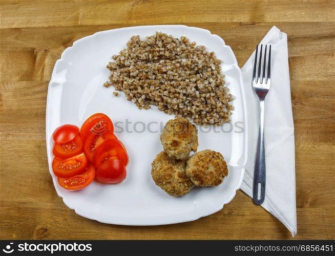 On a square plate is buckwheat porridge with meat cutlets and chopped tomato