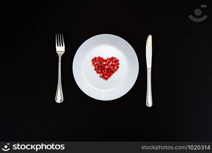 On a white plate from pomegranate seeds composite heart shape. Knife and fork next to the plate. Black background. Flat layer.
