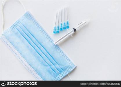 On a white table lies a syringe and injection needles against the background of a medical mask. The concept of vaccination and health. View from above. On a white table lies a syringe and injection needles against the background of a medical mask. The concept of vaccination and health. View from above.