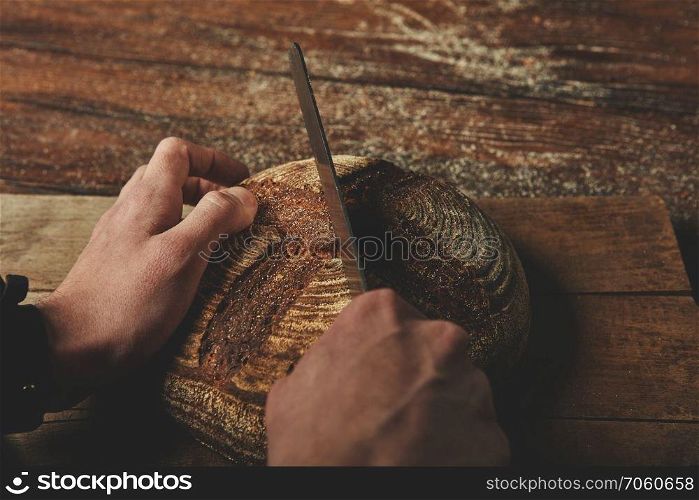 On a wooden brown background men&rsquo;s hands cut round rustic bread close-up. A man baker cuts bread