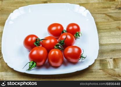 On a wooden table is a white plate with red small tomatoes