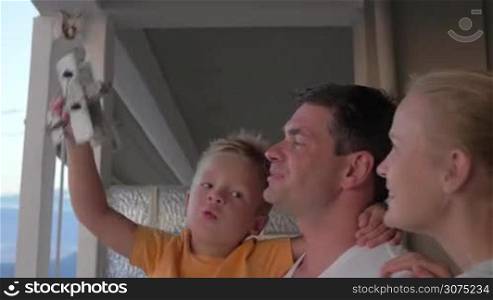 On balcony of house is mother and father with their son on hands. Son plays with a toy plane. Happy family together
