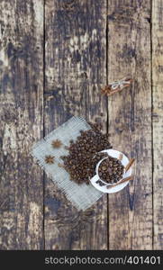 On the old wooden table is a white cup with grains of coffee. Grains of coffee are scattered on a linen napkin. The cinnamon is tied with a rope. Anise complements the aroma of coffee. Top view, vertical placement.. Grain coffee in a cup, which stands on a sackcloth from burlap. Cinnamon on a platter and tied with a rope. Anise stars complement the aroma of coffee. View from above.