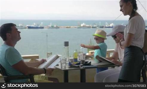 On the sea coast of city Perea, Greece at table in cafe sits a young family and ordered meals from the waitress