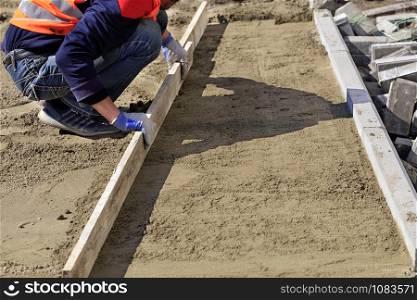 On the sidewalk, a worker cleans and levels the sandy platform with a wooden board, preparing the foundation for laying the paving slabs.. The worker clears and aligns the sand base with a wooden board for the subsequent laying of tiles on the sidewalk.