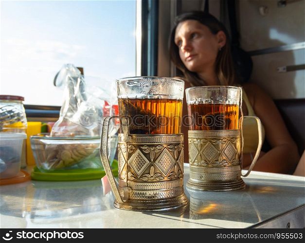 On the table in the compartment of the train are glasses with tea in metal cup holders, in the background the girl looks out the window
