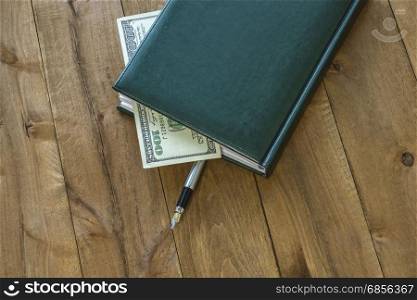 On the wooden surface is a diary with a pen and a part of the money bill is visible