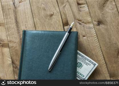 On the wooden surface is a diary with a pen and a part of the cash bill is visible
