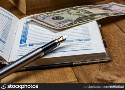 On the wooden surface is an open diary, a fountain pen and a money bill
