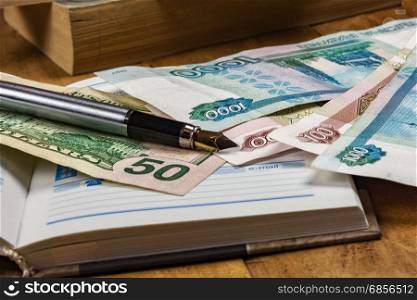 On the wooden surface is an open diary, money bills and fountain pen