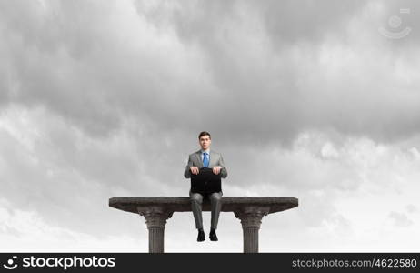 On top of world. Young businessman with briefcase sitting on top of building