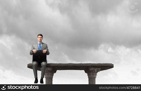 On top of world. Young businessman with briefcase sitting on top of building