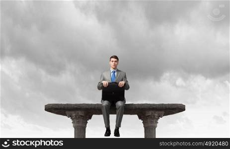 On top of world. Young businessman with briefcase sitting on top of building
