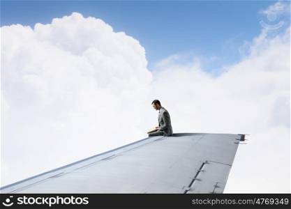 On wing of flying airplane. Young businessman sitting on edge of airplane wing with book in hands