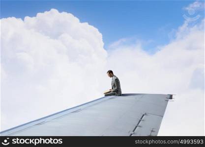 On wing of flying airplane. Young businessman sitting on edge of airplane wing with book in hands