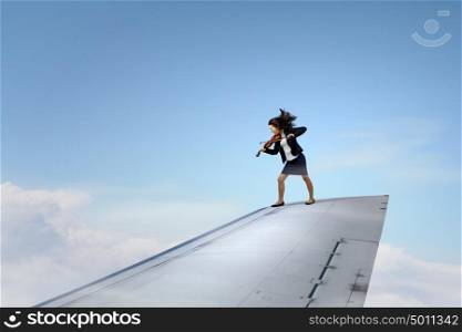 On wing of flying airplane. Young businesswoman standing on edge of airplane wing and play violin