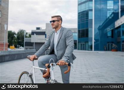 One businessman in sunglasses poses on bicycle at the office building in downtown. Business person riding on eco transport on city street