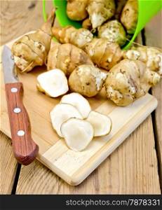 One cut and a few whole tubers of Jerusalem artichoke with a green bucket, knife on a wooden boards background