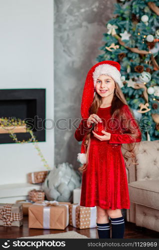 One cute cheerful girl santa hat near Christmas tree. Holds red christmas ball. Merry Christmas.. One cute cheerful girl santa hat near Christmas tree. Holds red christmas ball.