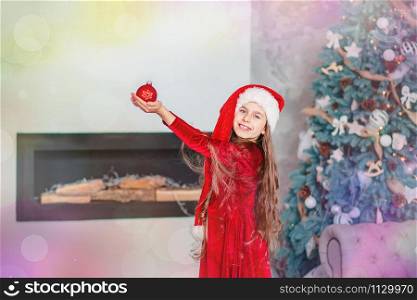 One cute cheerful girl santa hat near Christmas tree. Holds red christmas ball. Merry Christmas.. One cute cheerful girl santa hat near Christmas tree. Holds red christmas ball.