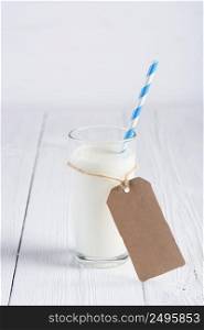 One glass of milk with blue stripped paper straw and blank paper tag on rope on white wooden table still life