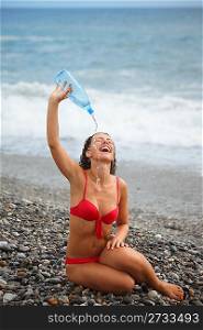 one joyful woman wearing swimsuit with bottle is sitting near water on sea coast