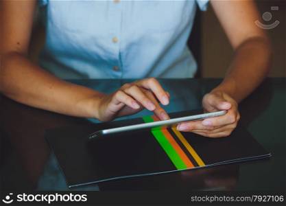 One light-skinned hand belonging to a woman holds a large tablet while the other hand uses a pointer finger to access something on the tablets touch screen display. The tablet has a gray case and a glass screen. The woman in blue shirt.. Woman working on digital tablet